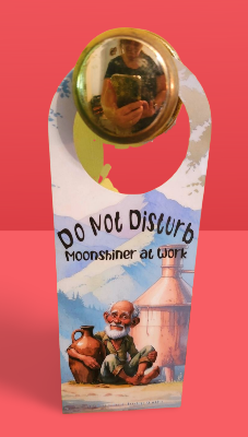 Door hanger with illustration of a moonshiner and text 'Do Not Disturb Moonshiner at Work' on a red background