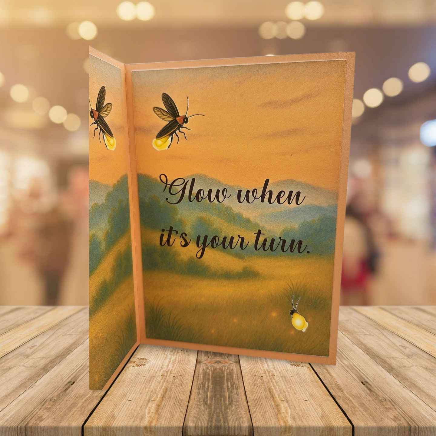 Greeting card with glowing insects and 'Glow when it's your turn' text on a wooden surface.