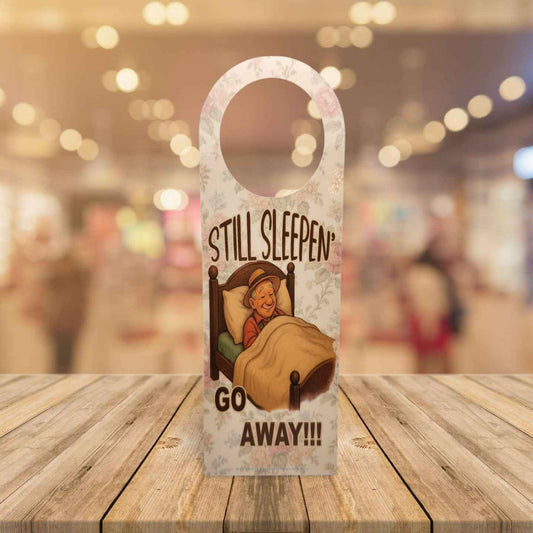 Door hanger with cartoon character and text 'STILL SLEEPEN GO AWAY!!!' on a wooden surface.