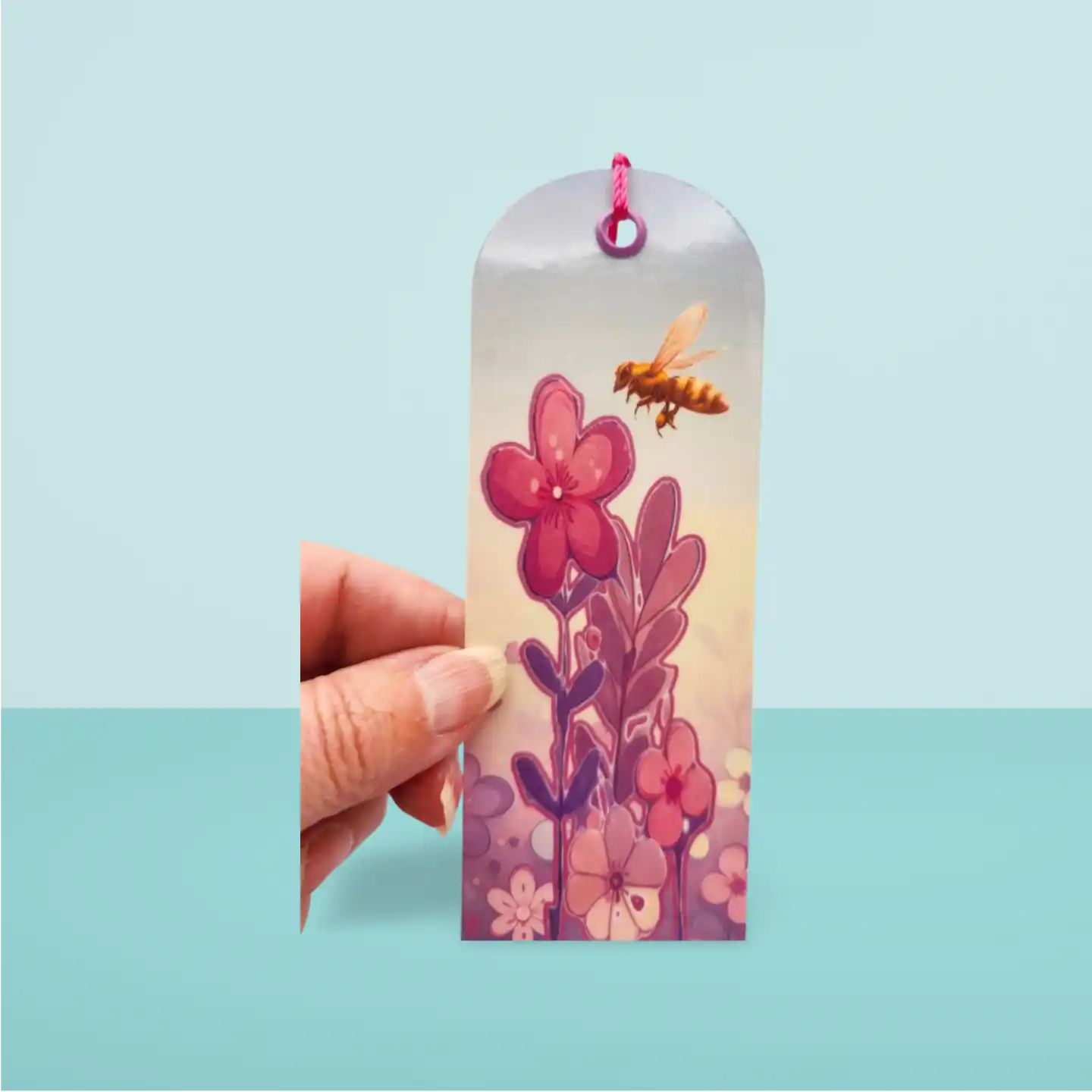 Hand holding a bookmark with floral design and bee illustration against a light blue background