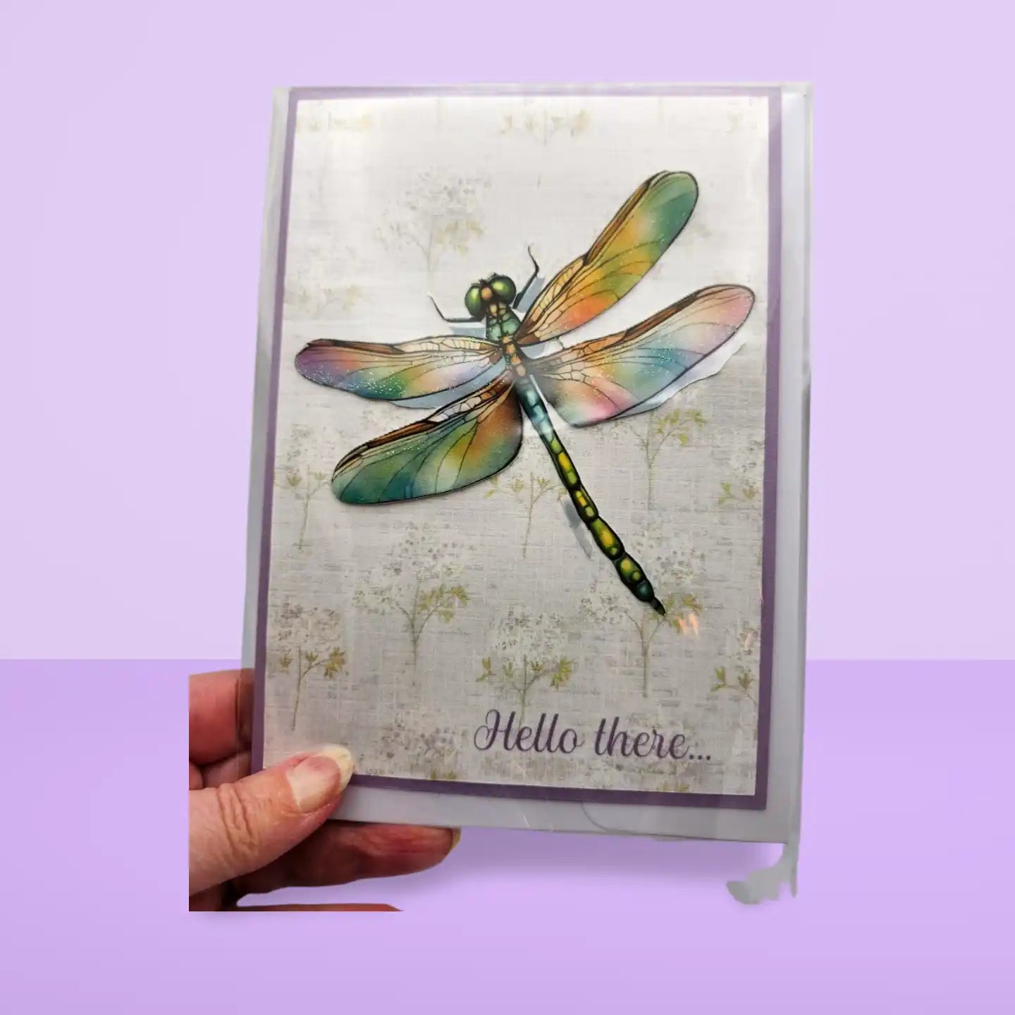 Card with a colorful dragonfly design and 'Hello there...' text, held against a purple background.