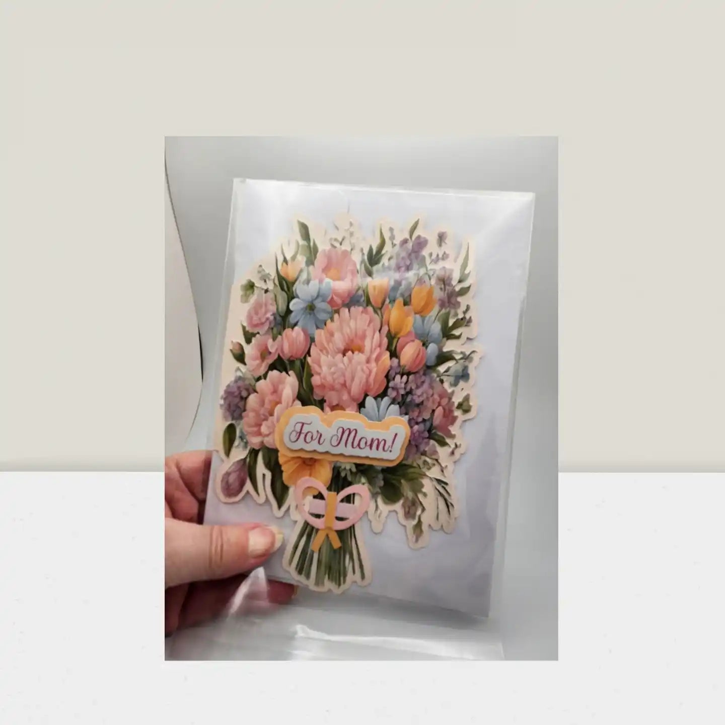 Greeting card with floral design and 'For Mom!' text held by a hand on a white background