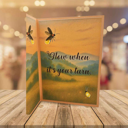 Greeting card with glowing insects and 'Glow when it's your turn' text on a wooden surface.