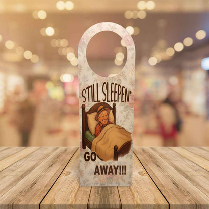Door hanger with cartoon character and text 'STILL SLEEPEN GO AWAY!!!' on a wooden surface.
