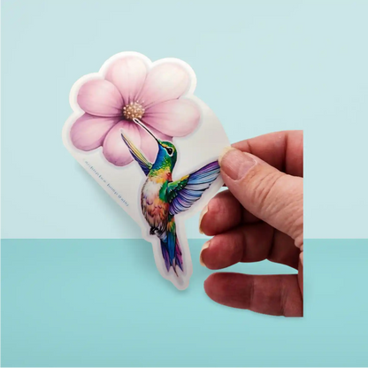 Hand holding a colorful hummingbird and flower sticker against a light blue background
