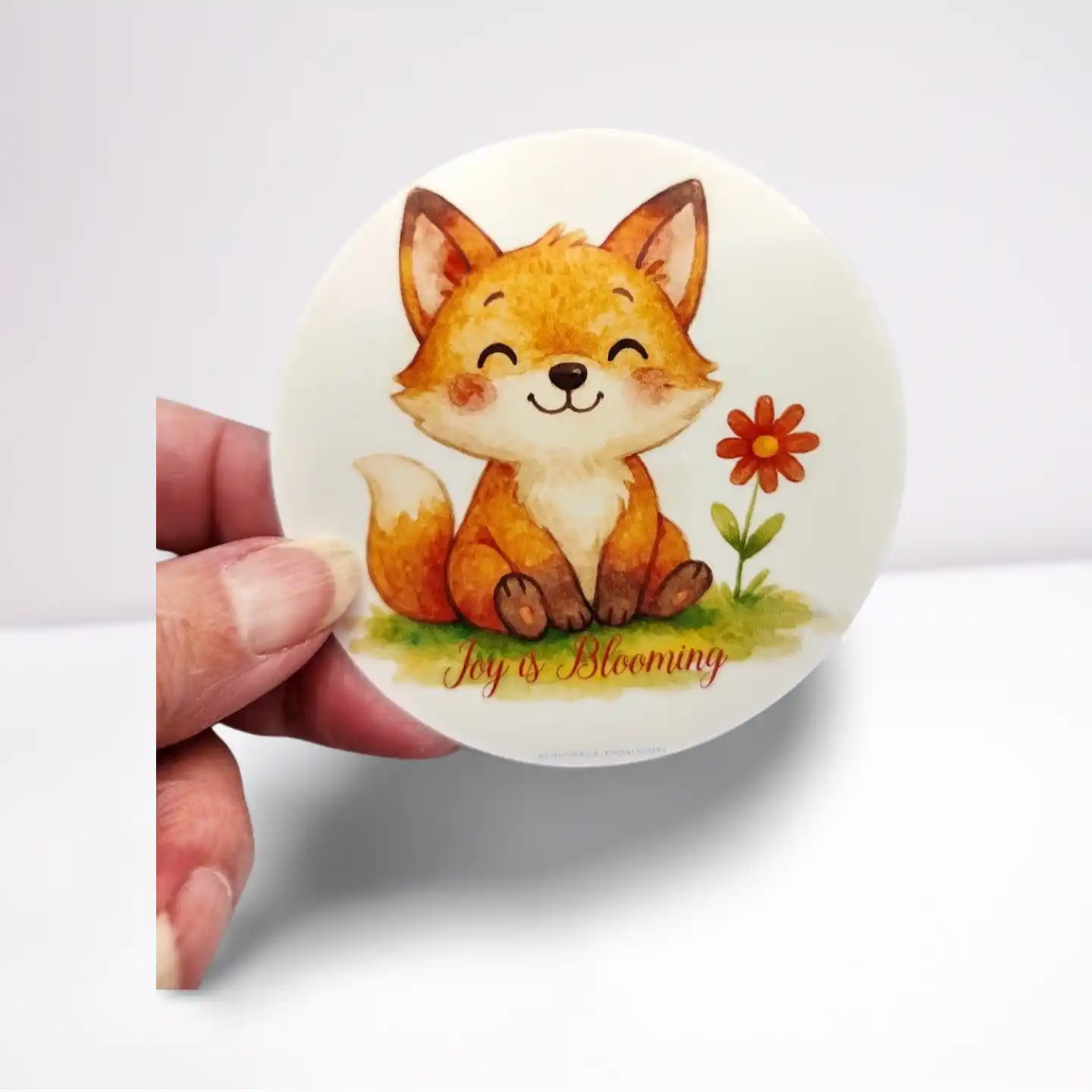 Round sticker with a cartoon fox and flower design held by a hand on a white background