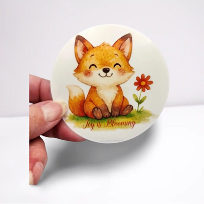 Round sticker with a cartoon fox and flower design held by a hand on a white background