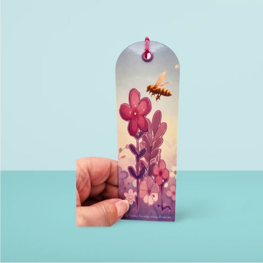 Hand holding a bookmark with floral design and bee illustration on a light blue background