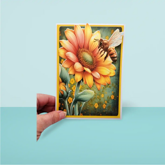 Hand holding a card with a sunflower and bee design against a light blue background