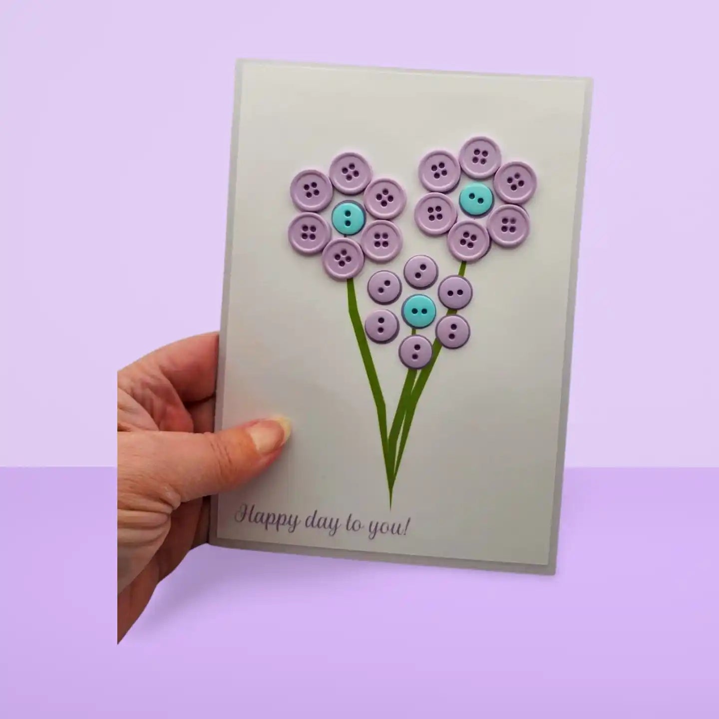 Handmade card with button flower design held by a hand on a purple background