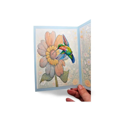 Greeting card with a colorful illustration of a hummingbird on a flower, held open by a hand against a white background.