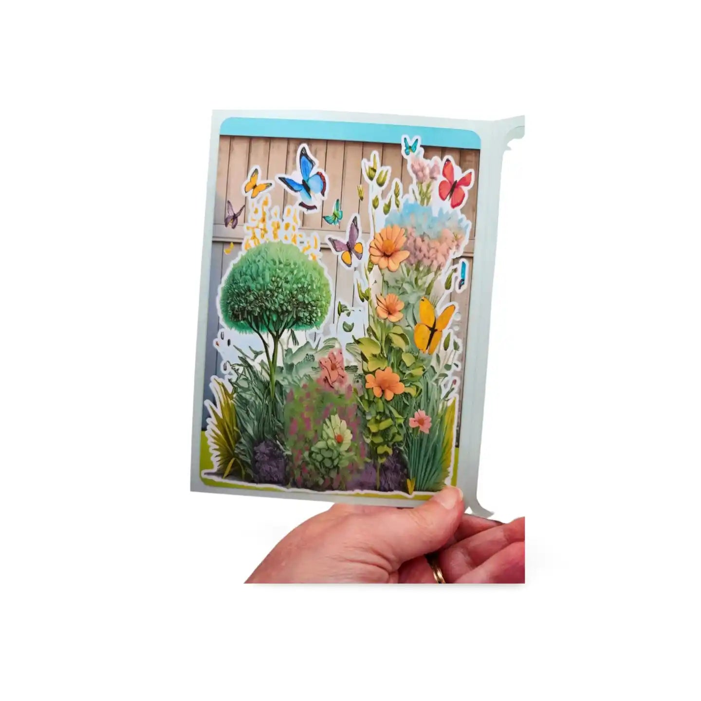 Hand holding a 3D garden scene card with flowers and butterflies on a white background