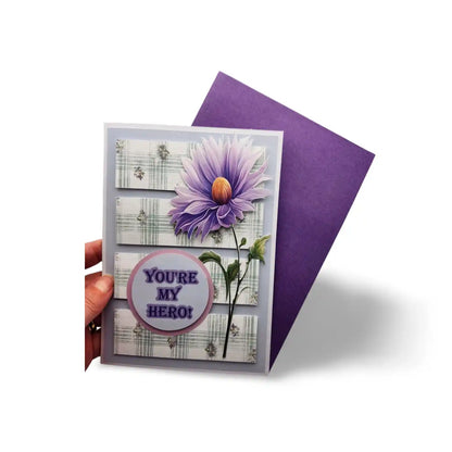 Greeting card with purple flower design and 'You're My Hero!' text, held by a hand.