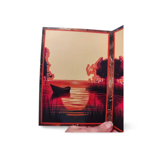 Foldable card with a sunset over water design held open by a hand on a white background