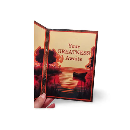 Card with a scenic design and 'Your GREATNESS Awaits' text, held open by a hand.
