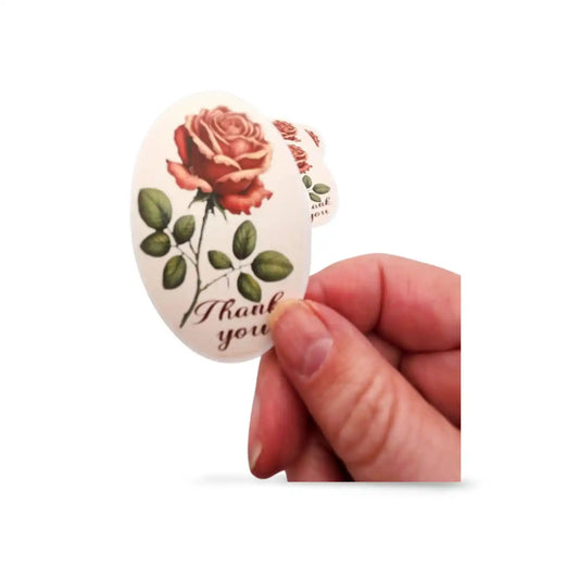 Hand holding a round sticker with a rose design and 'Thank you' text on a white background