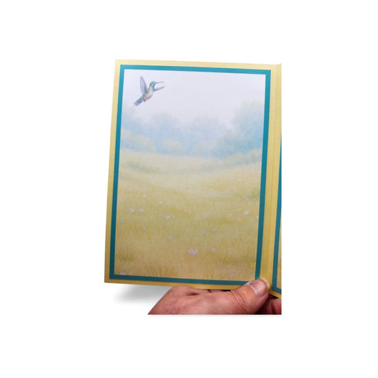 Hand holding a card with a landscape of a field and bird in the sky.