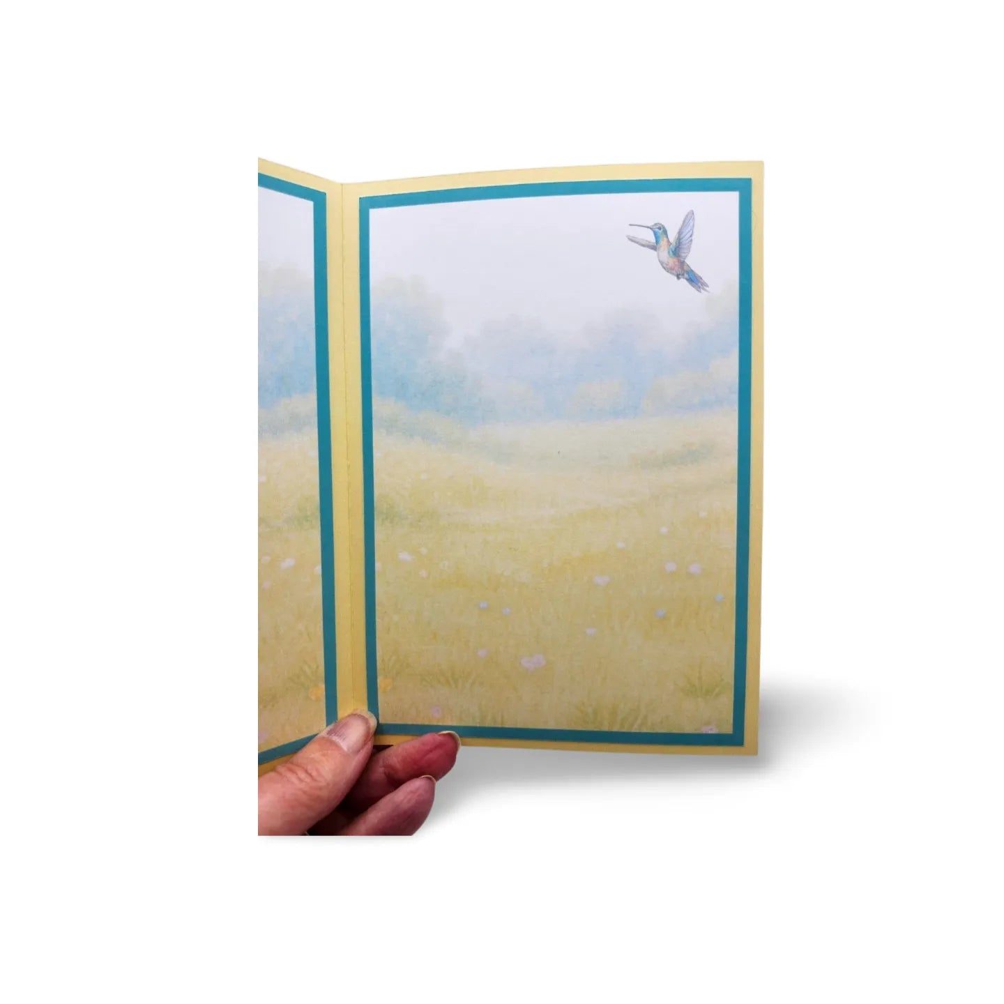 Hand holding a greeting card with a scenic illustration of a field and bird.