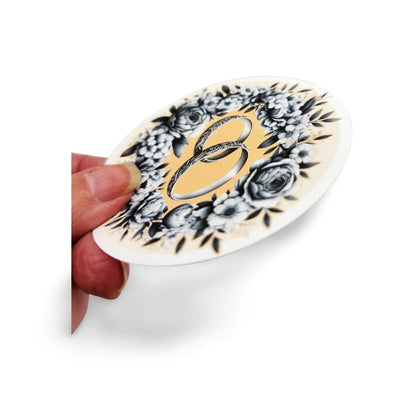 Decorative oval sticker with floral design and letter 'B' held by a hand on a white background