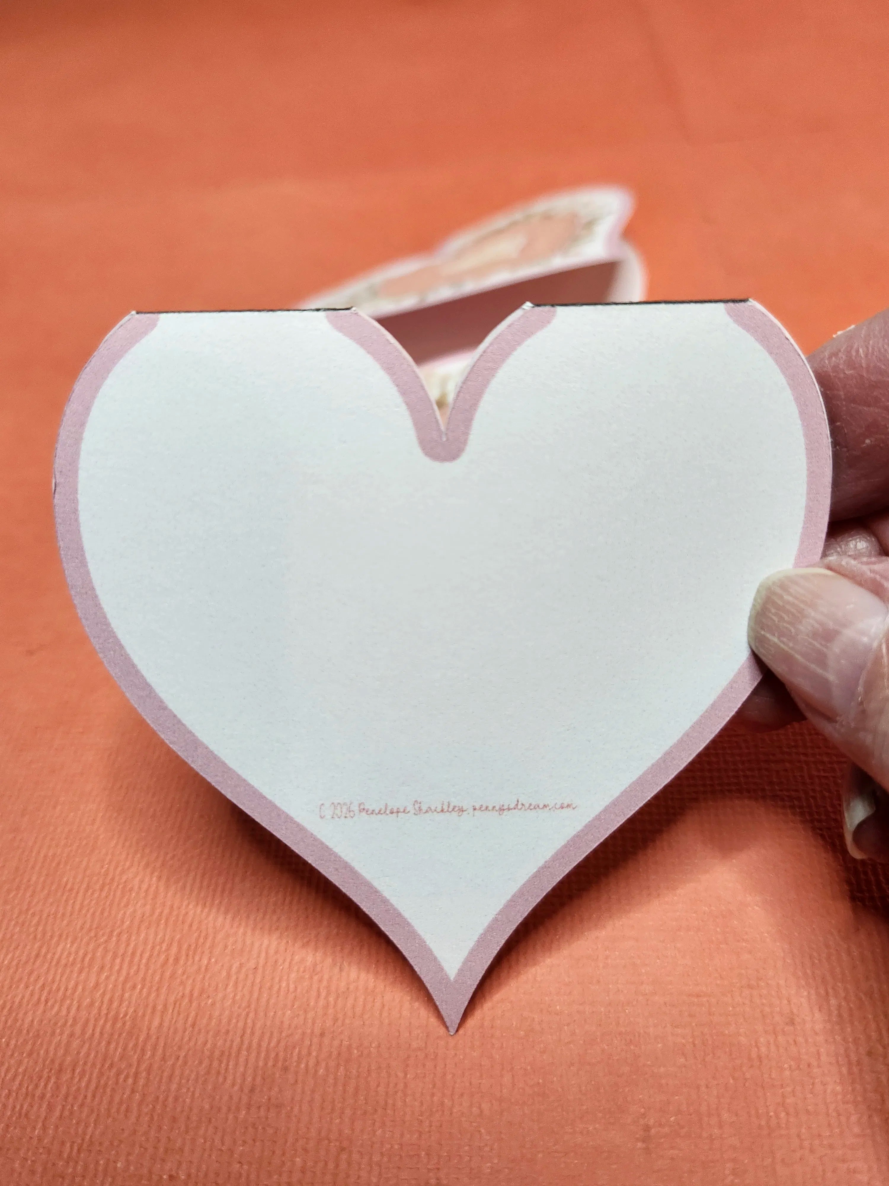 Heart-shaped white card with pink border held by a hand on an orange background