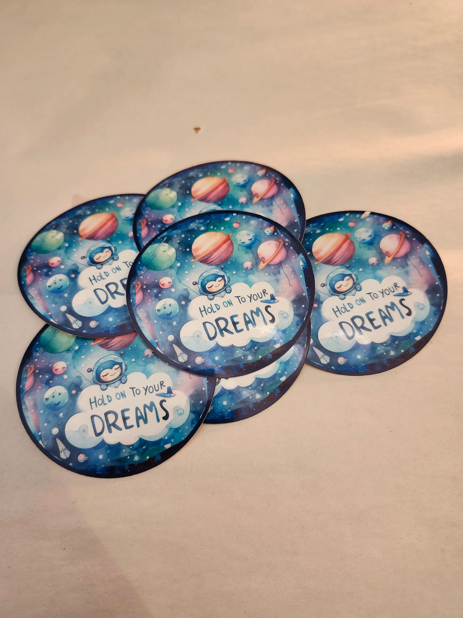 Five circular stickers with space-themed design and 'Hold on to your dreams' text on a beige background.