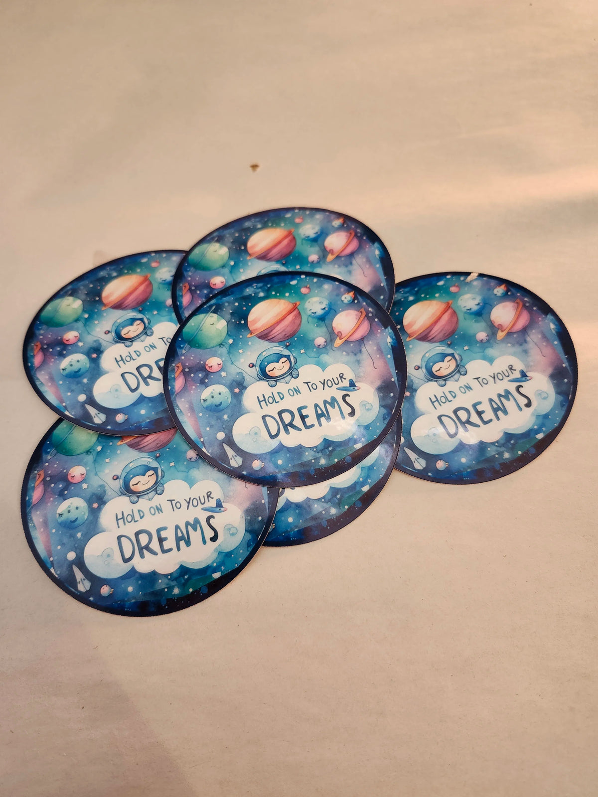 Five circular stickers with space-themed design and 'Hold on to your dreams' text on a beige background.