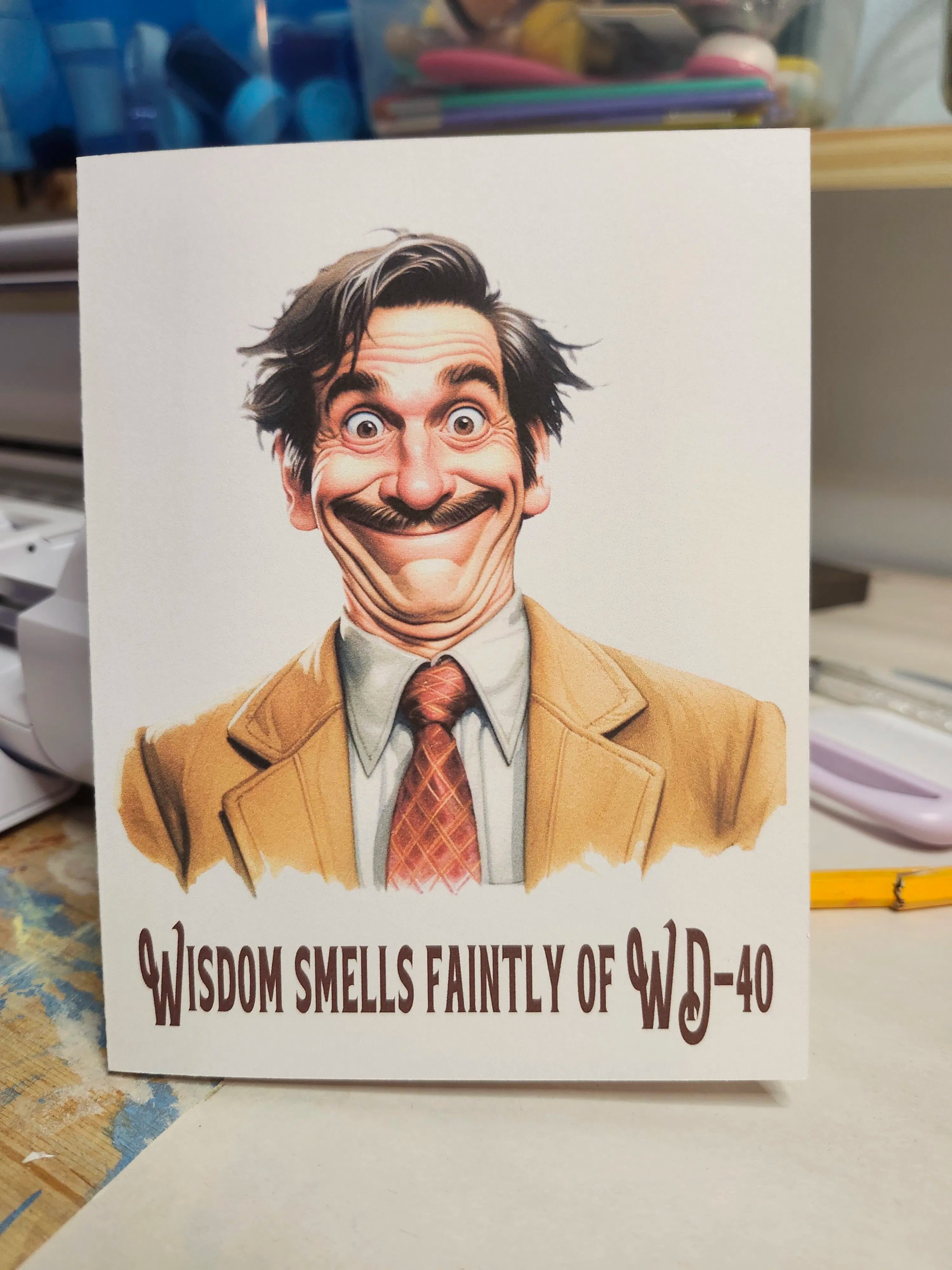 Card with a humorous illustration of a man and text 'Wisdom smells faintly of WD-40' on a desk background.