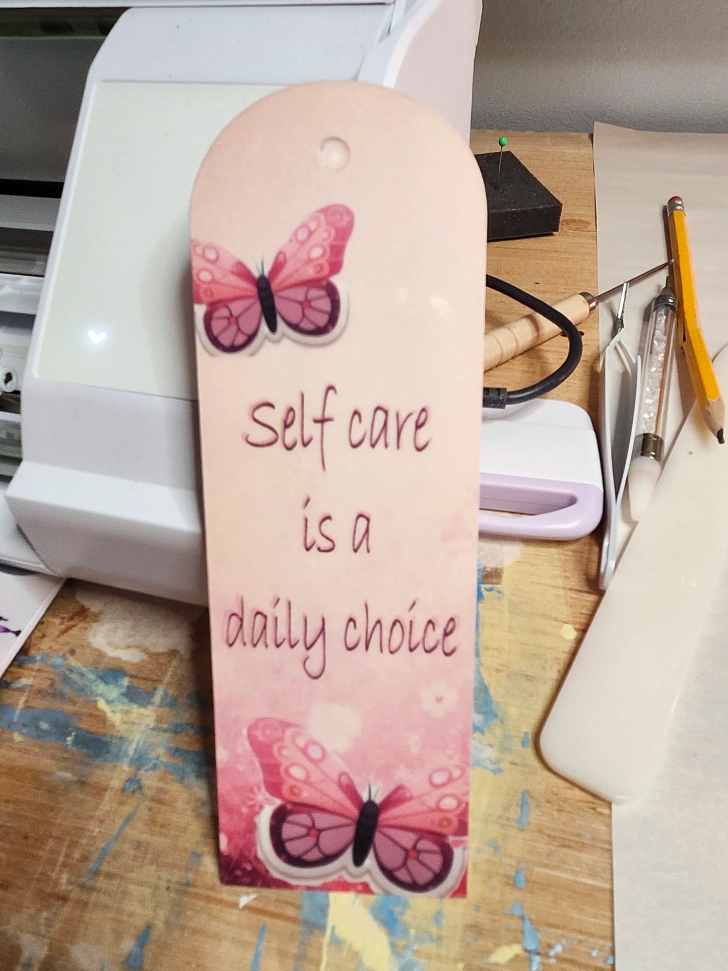 Bookmark with butterfly design and text 'Self care is a daily choice' on a desk.