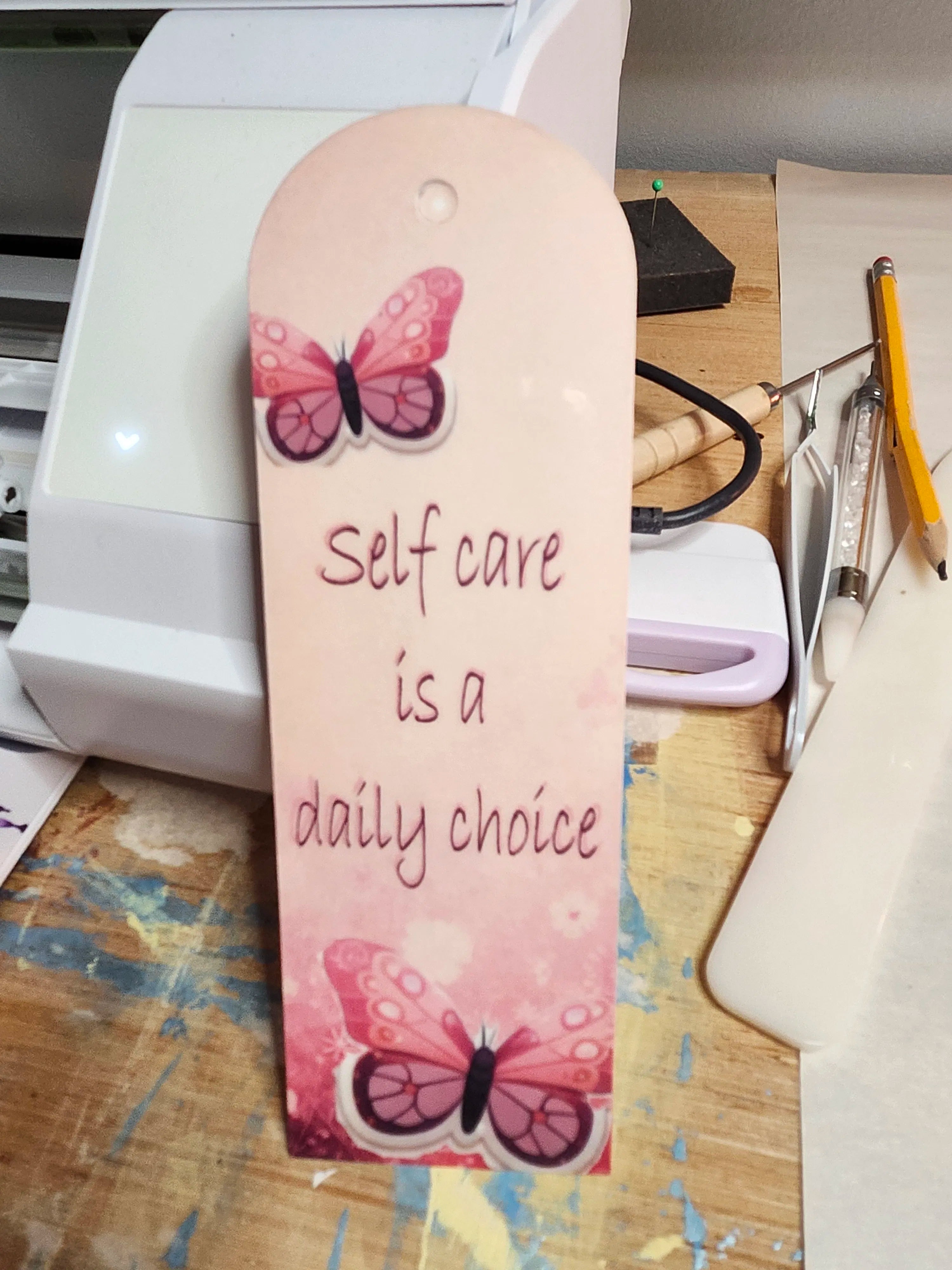Bookmark with butterfly design and text 'Self care is a daily choice' on a desk.
