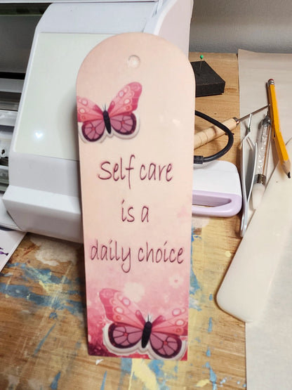 Bookmark with butterfly design and text 'Self care is a daily choice' on a desk.