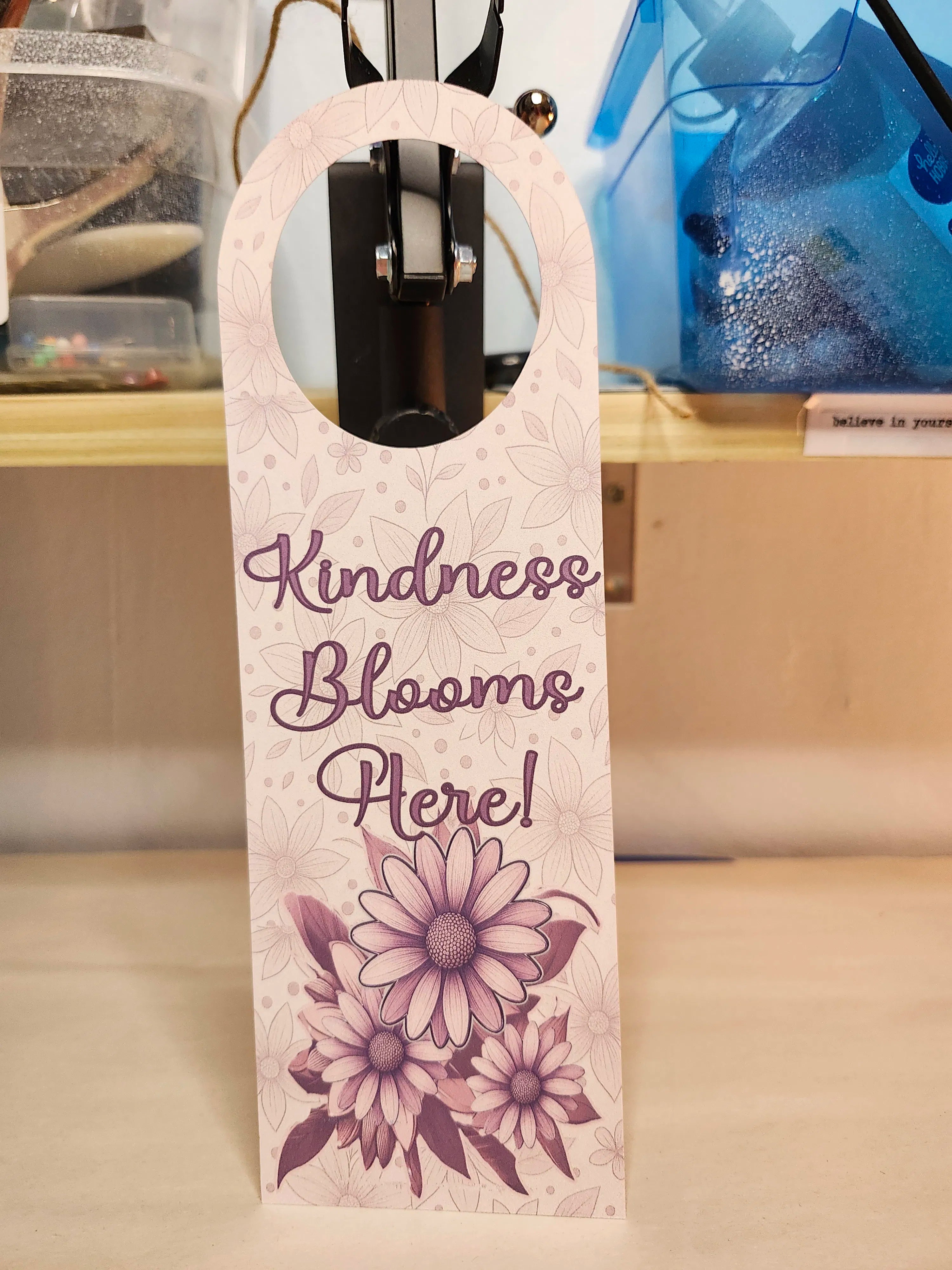Decorative bottle tag with 'Kindness Blooms Here!' text and floral design on a blurred background.