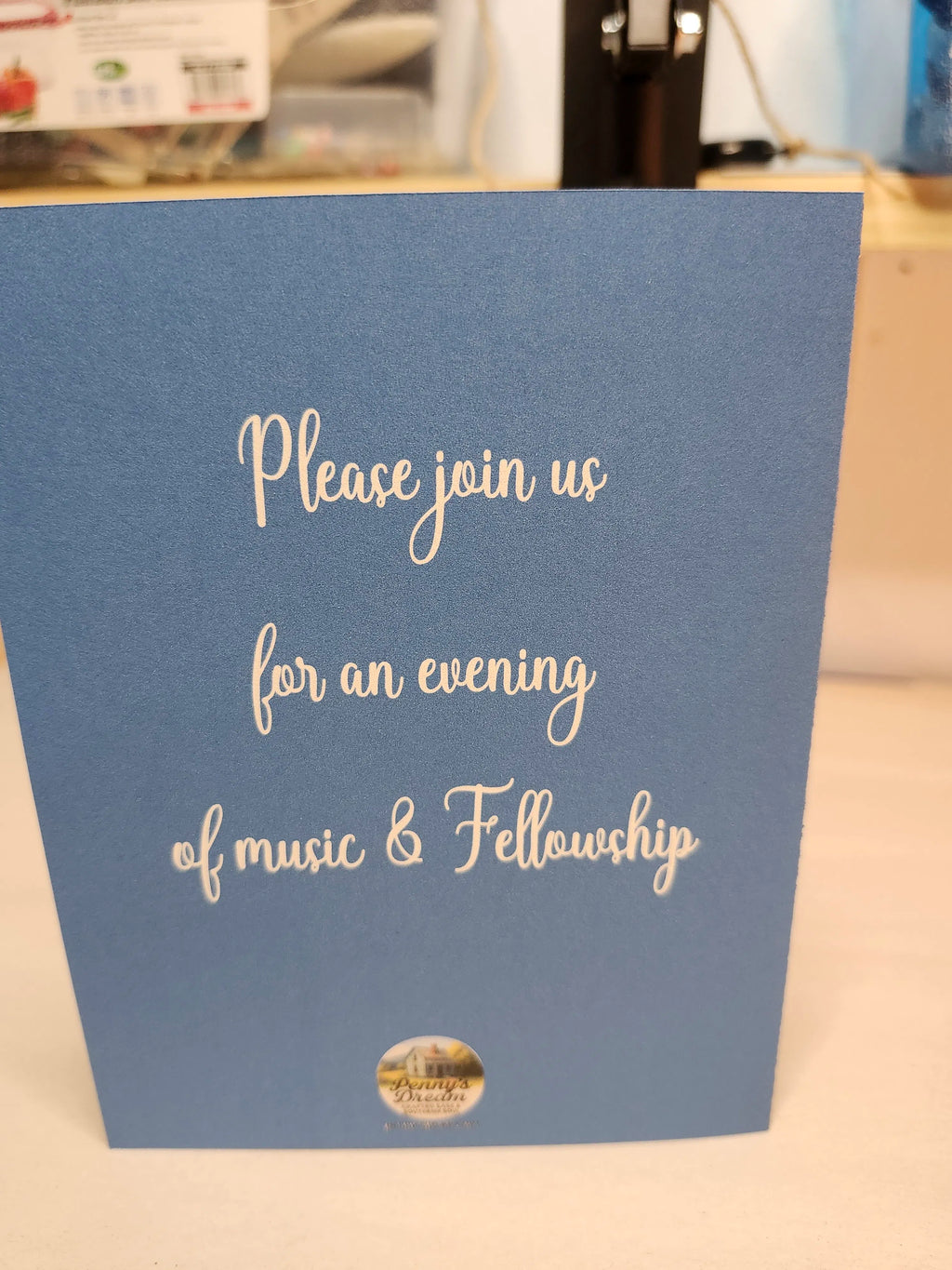 Blue card with white text inviting to an evening of music and fellowship, on a blurred indoor background.