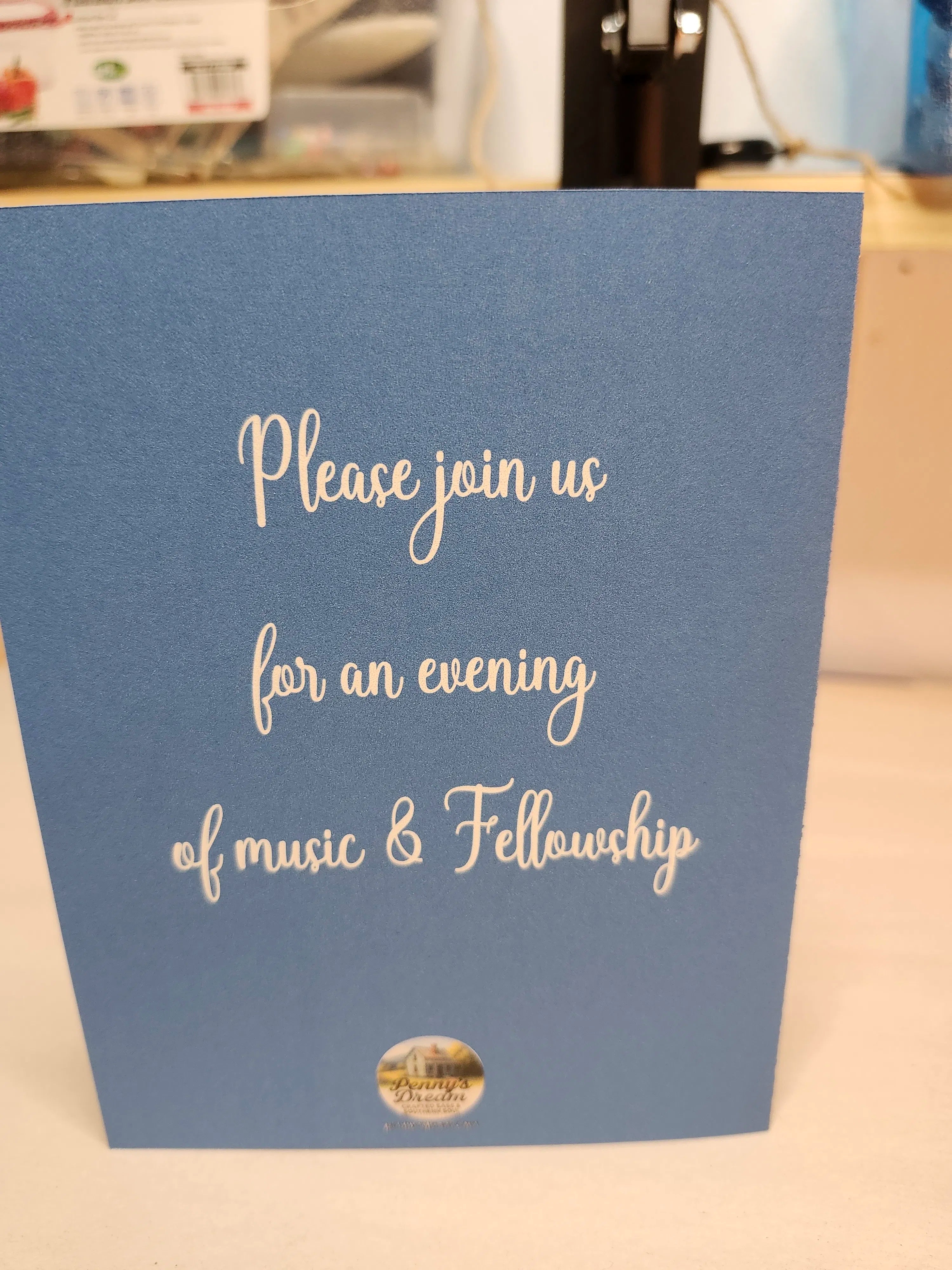 Blue card with white text inviting to an evening of music and fellowship, on a blurred indoor background.