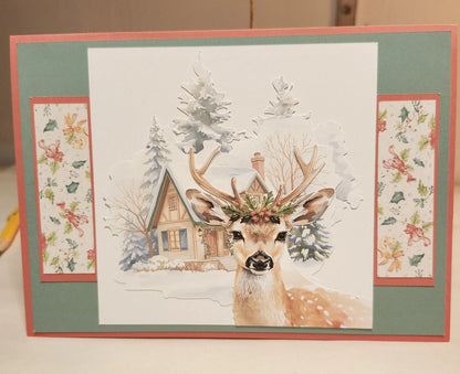 Card with a deer illustration and snowy cabin in a decorative frame.