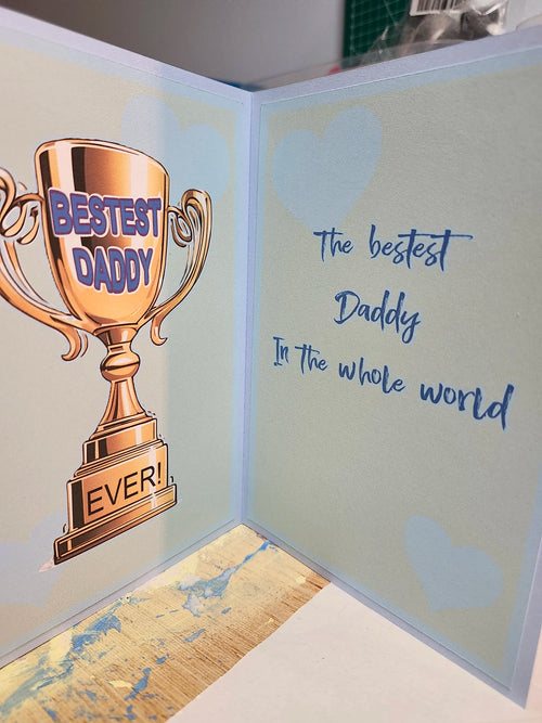 Greeting card with a trophy-shaped cutout and text 'Bestest Daddy Ever!' on a light blue background.