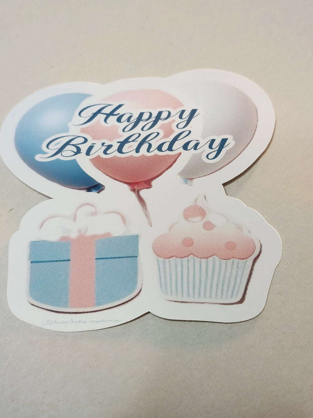 Sticker with 'Happy Birthday' text, balloons, gift box, and cupcake on a beige background