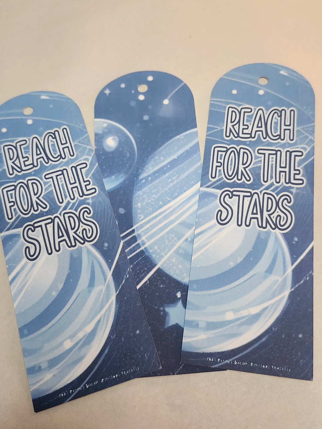 Two blue bookmarks with 'Reach for the Stars' text and celestial design on a beige background.