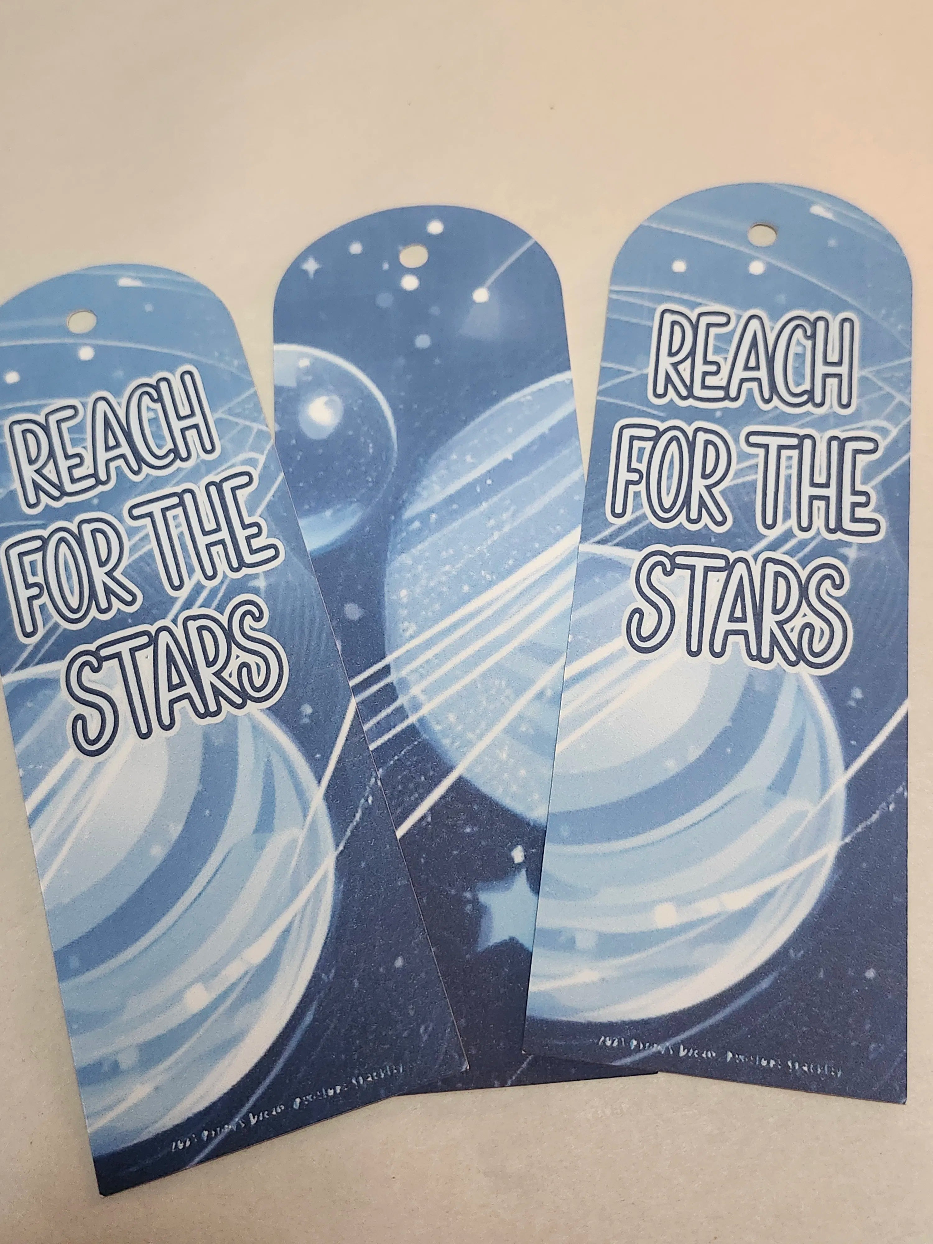 Two blue bookmarks with 'Reach for the Stars' text and celestial design on a beige background.