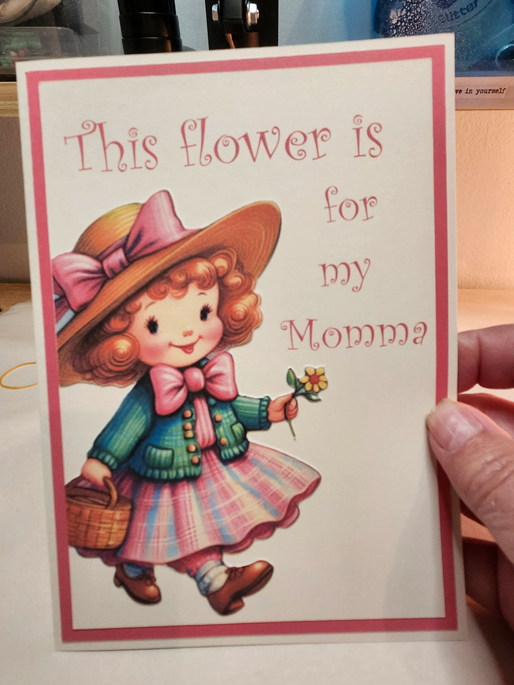 Card with illustration of a girl holding a flower and text 'This flower is for my Momma' held by a person.