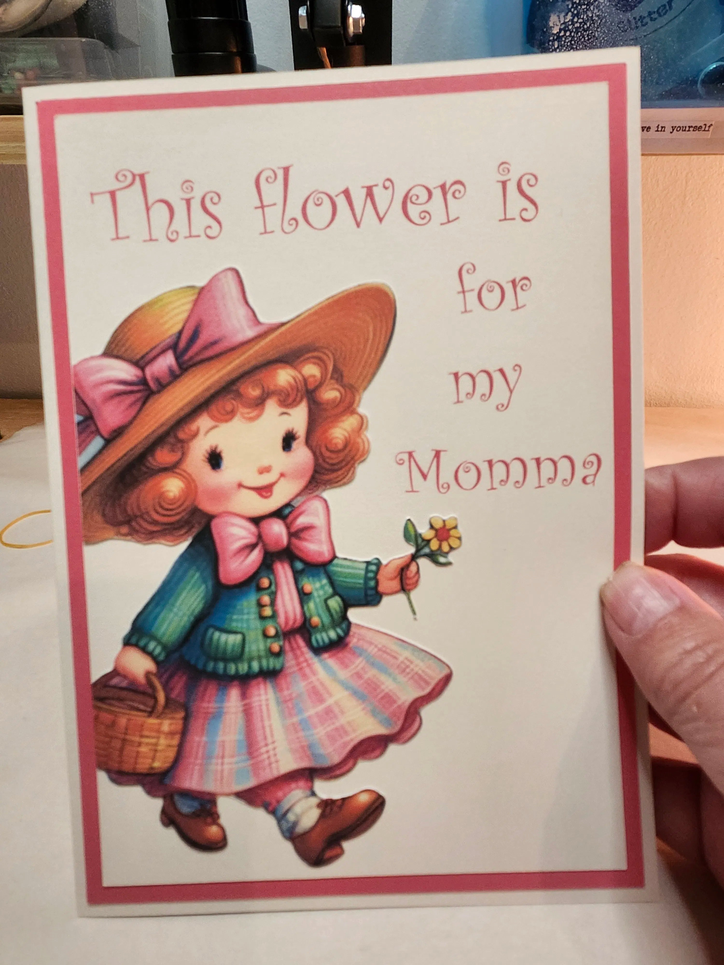 Card with illustration of a girl holding a flower and text 'This flower is for my Momma' held by a person.