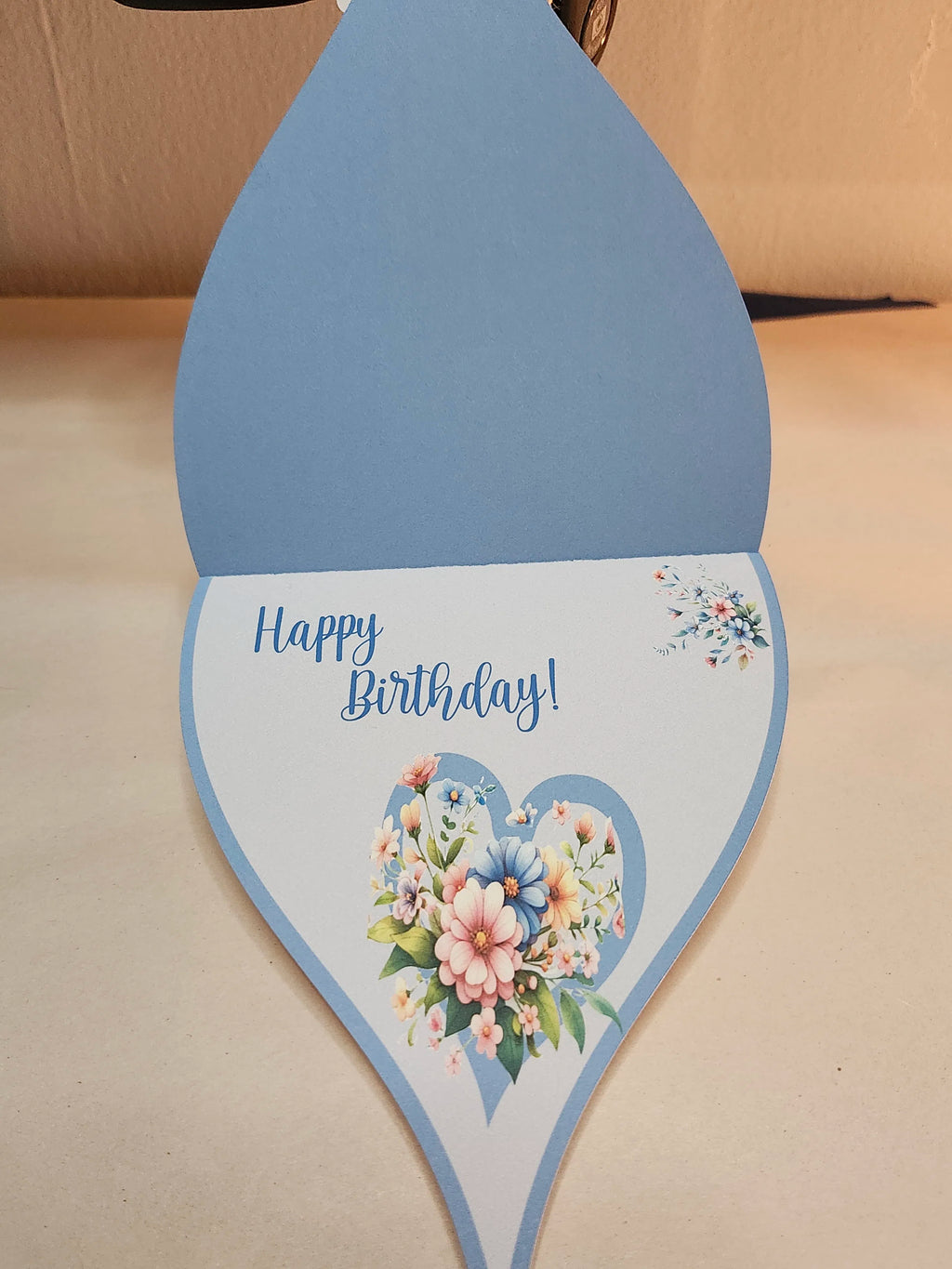 Teardrop-shaped birthday card with floral design and 'Happy Birthday!' text on a beige surface.