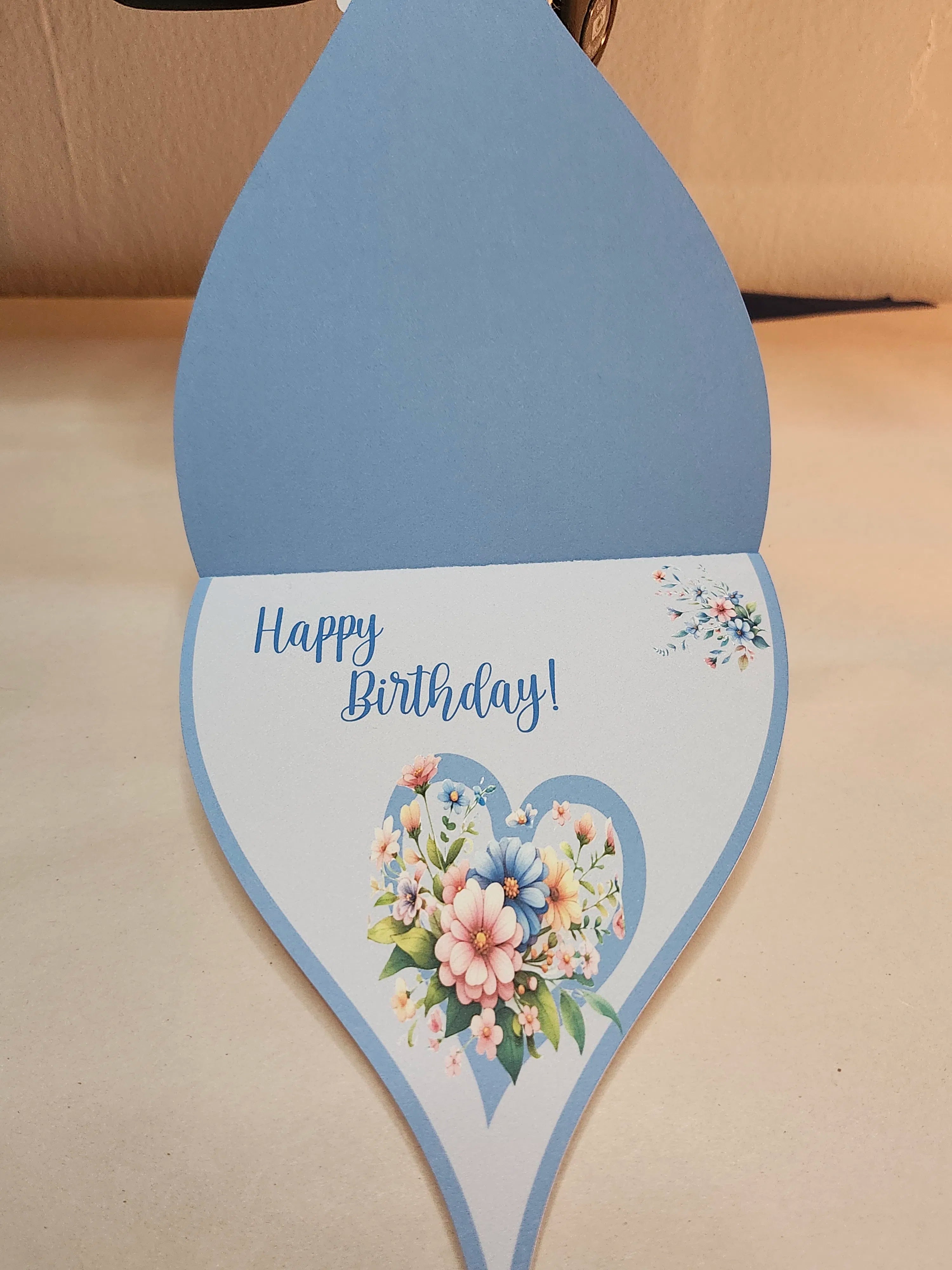 Teardrop-shaped birthday card with floral design and 'Happy Birthday!' text on a beige surface.