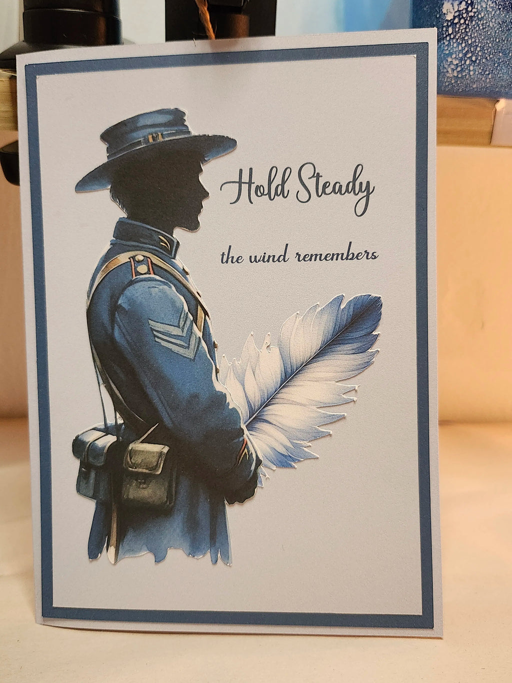 Greeting card with a silhouette of a person in uniform, a feather, and text 'Hold Steady the wind remembers' on a light background.