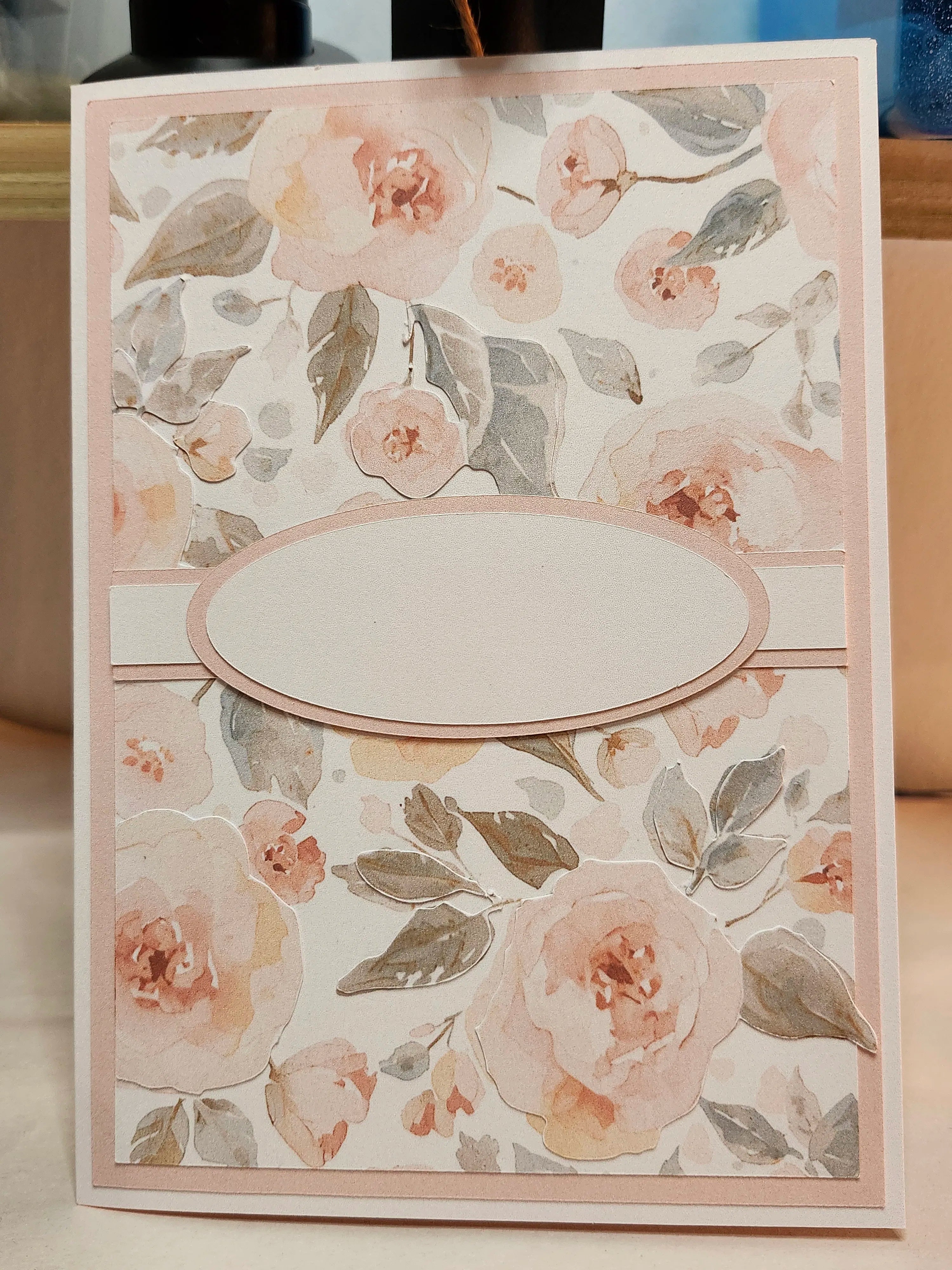 A handmade greeting card featuring a watercolor floral pattern with pink and beige roses and gray-green leaves, complete with a central blank oval label.