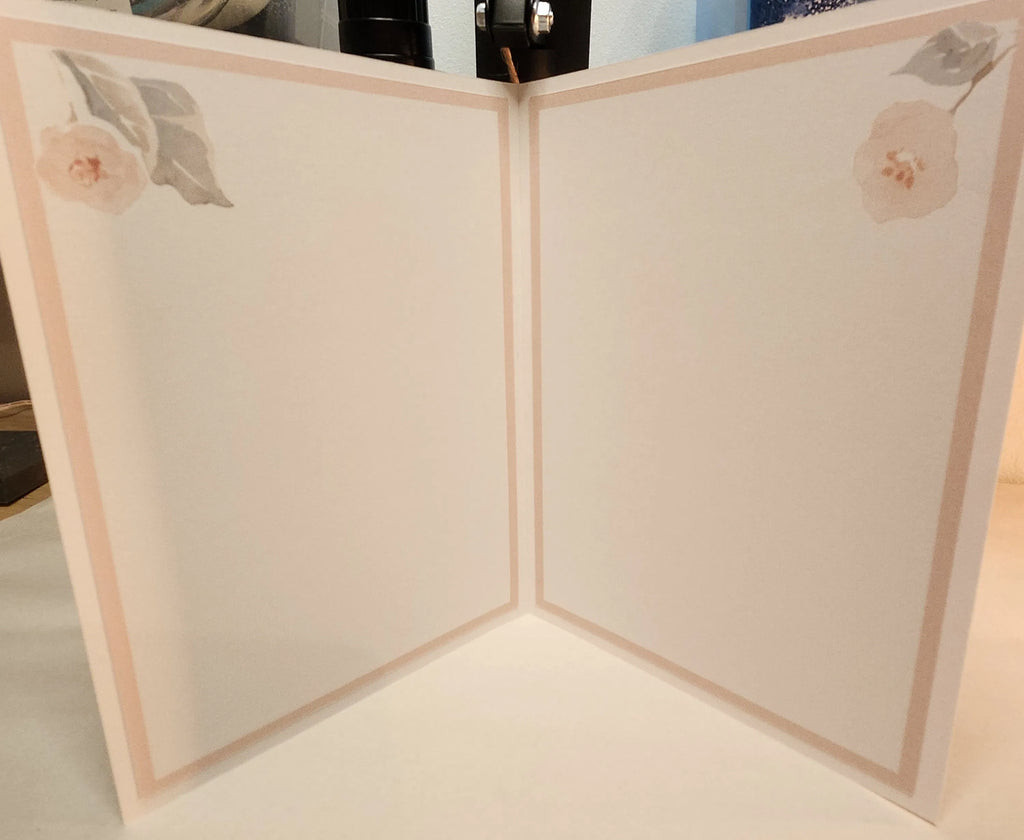 A cream-colored folding card or book shown open, featuring a delicate peach border and soft pink floral illustrations in the top corners.