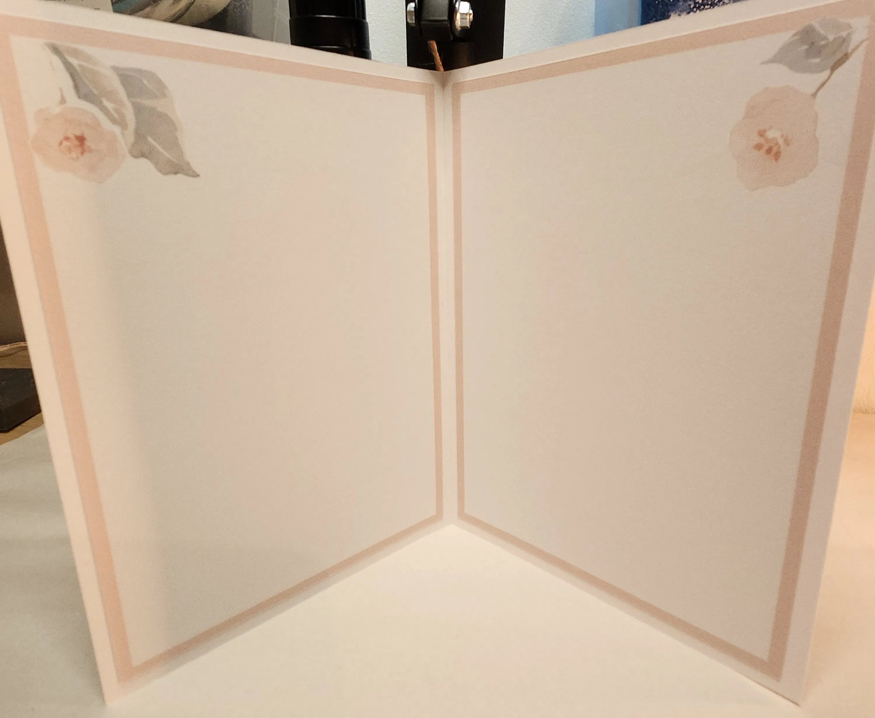 A cream-colored folding card or book shown open, featuring a delicate peach border and soft pink floral illustrations in the top corners.