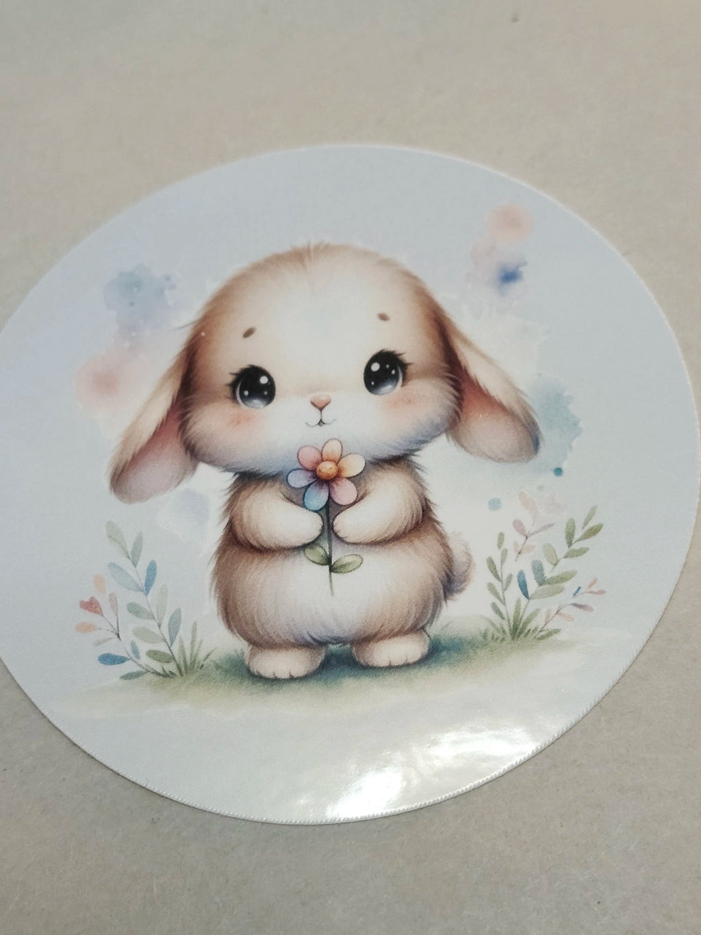 A round sticker featuring a cute watercolor-style illustration of a fluffy cream-colored bunny holding a small flower, set against a neutral beige background.