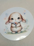 A round sticker featuring a cute watercolor-style illustration of a fluffy cream-colored bunny holding a small flower, set against a neutral beige background.