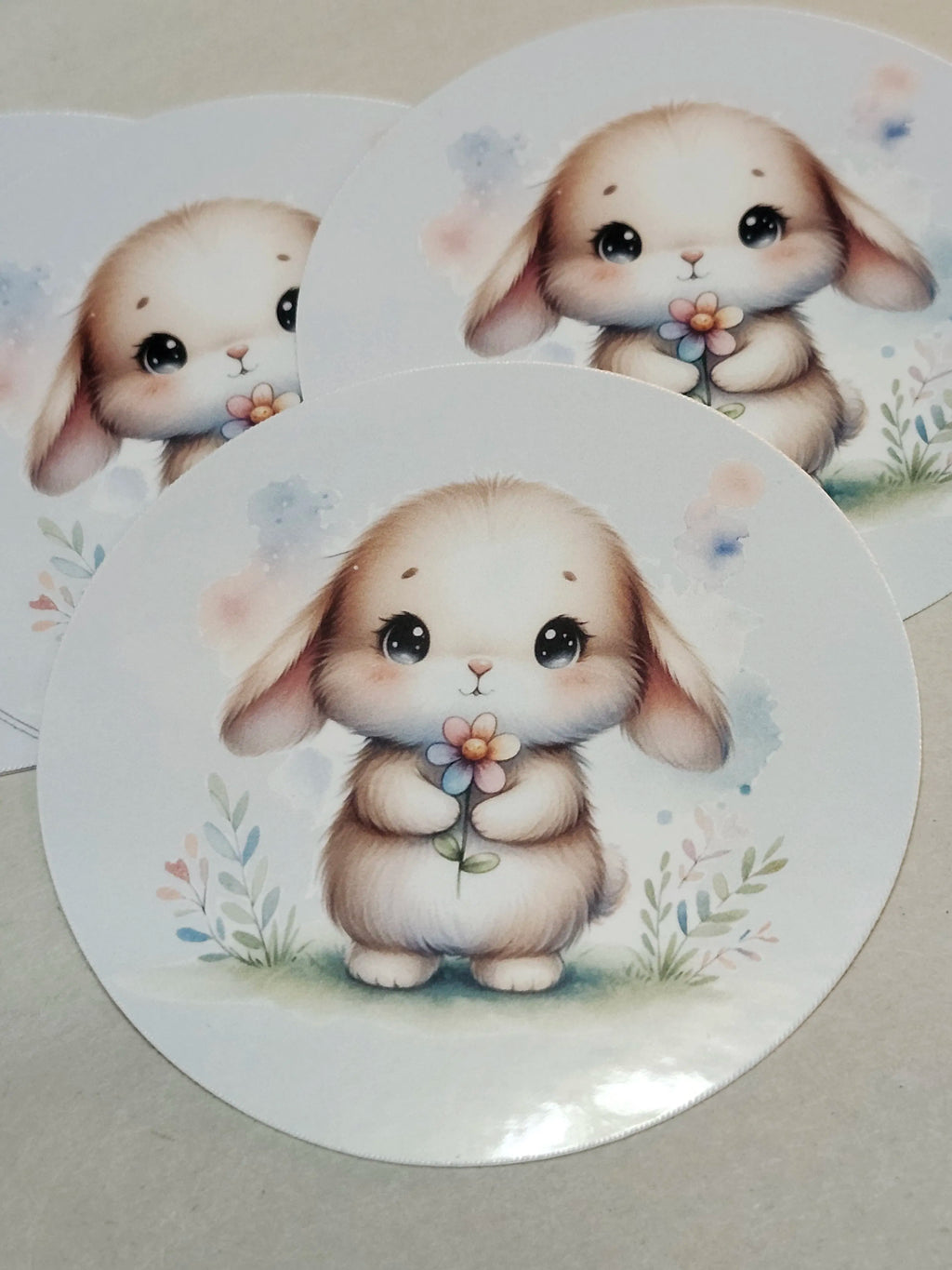 Three round white stickers featuring a cute cartoon bunny illustration holding a small flower, arranged on a light beige surface.