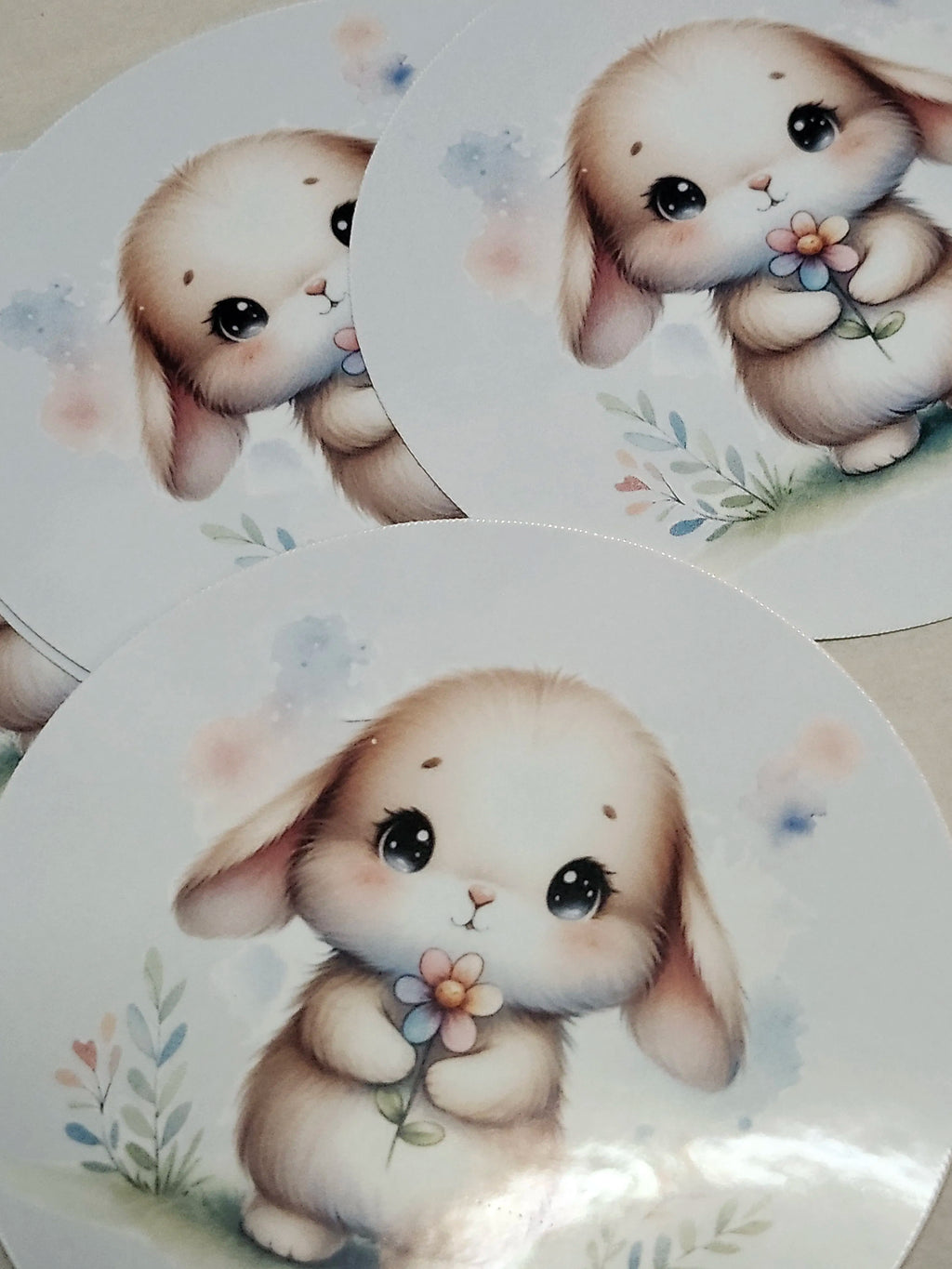 Three round stickers featuring a cute cartoon bunny illustration holding a small flower, shown overlapping on a light surface.